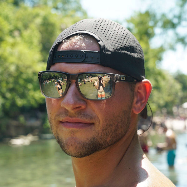 Man wearing black mens trucker hats with adjustable snapback and sunglasses outdoors near water and trees