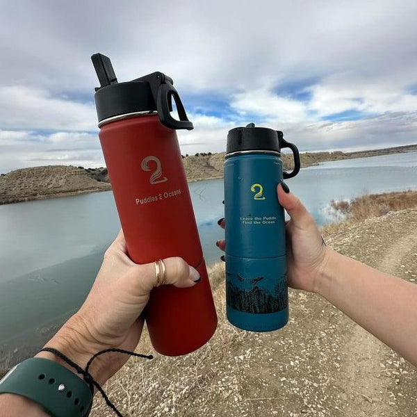 Two kids water bottles with storage compartments in red and blue held outdoors by water and rocky terrain