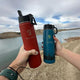 Two kids water bottles with storage compartments in red and blue held outdoors by water and rocky terrain