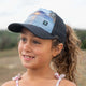 Youth girls wearing Seas the Day Girl Trucker Hat with high ponytail opening and coastal design