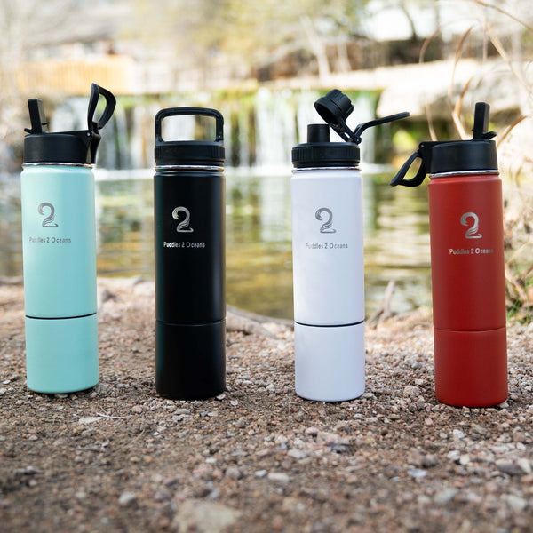 Set of four Sip N Seek 26oz kids water bottles in blue, black, white, and red with insulated stainless steel, bottom storage compartment, and multiple BPA-free lids on outdoor gravel background