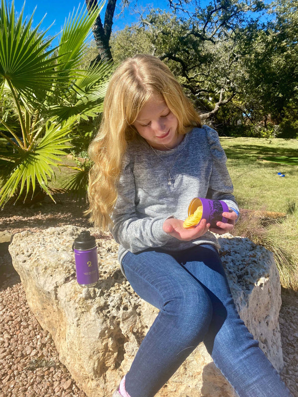 Child sitting outdoors on a rock using a purple kids water bottle with removable bottom storage compartment for snacks